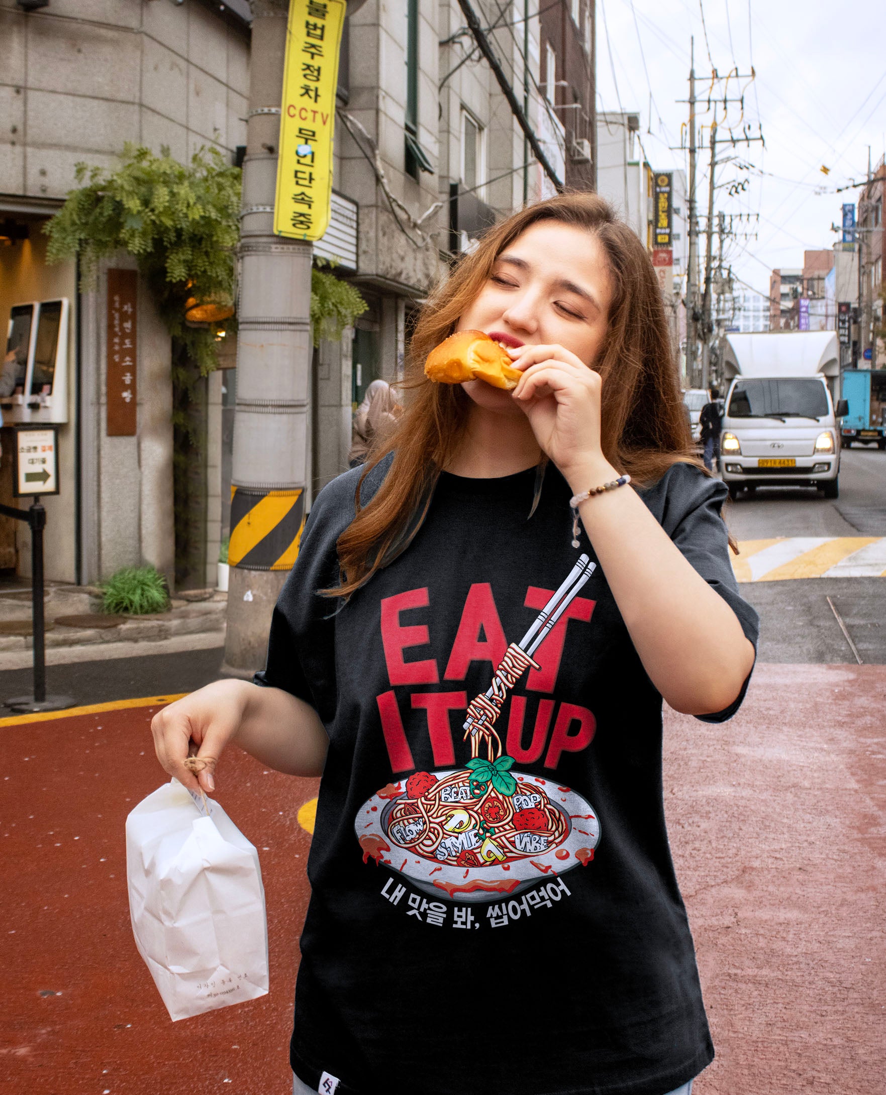 Woman eating a snack on a city street wearing a black t-shirt with a graphic design.